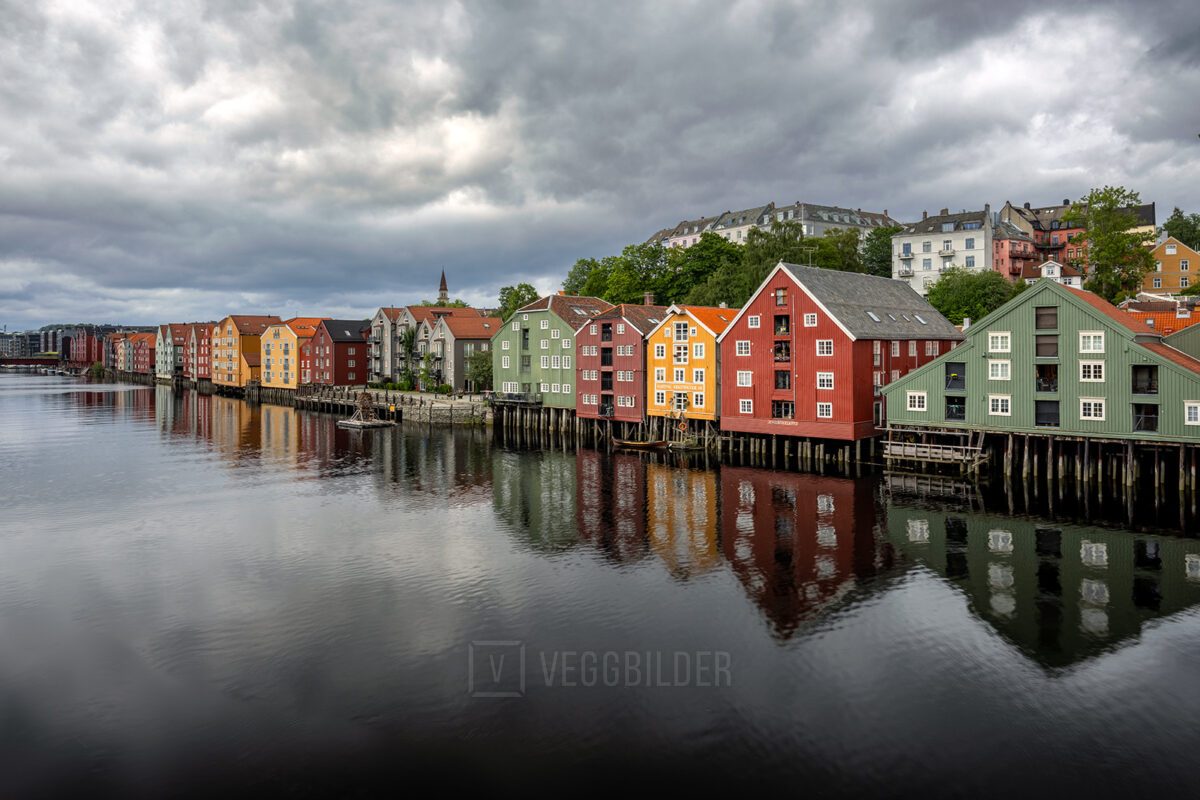 Fargerike trebygninger langs Nidelva i Trondheim på en overskyet dag, med speilblankt vann som gir en vakker refleksjon., fotokunst veggbilde / plakat av Eirik Sørstrømmen