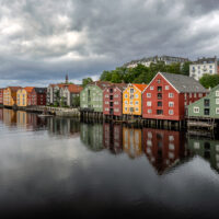 Fargerike trebygninger langs Nidelva i Trondheim på en overskyet dag, med speilblankt vann som gir en vakker refleksjon., fotokunst veggbilde / plakat av Eirik Sørstrømmen